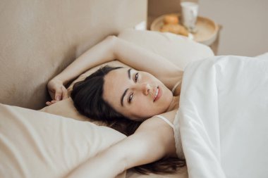 Portrait of young brunette woman waking up, lying on bed stretching arms and looking at camera with sensual look. Horizontal