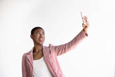 Smiling Black Young woman with buzz cut hairstyle in pink blazer taking selfie showing tongue using smartphone while standing against white wall. Horizontal