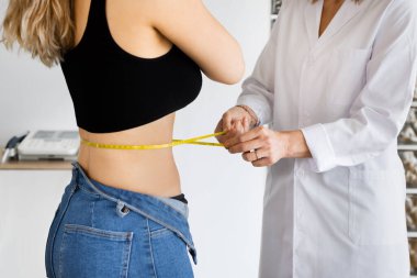 Side view close up of anonymous dietitian with flexible tape measuring waist of woman with crossed arms in office. Horizontal