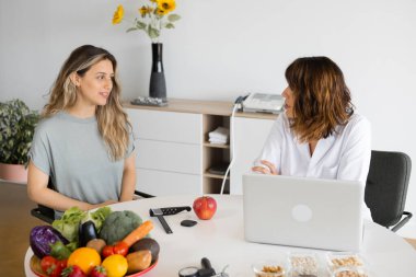 Female Nutritionist Advising A Client In Weight Loss Clinic. Mature nutritionist sitting at table and listening to patient in office, with laptop and fresh vegetables on desk.