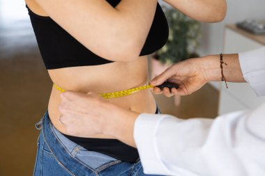 Close up of hands of Anonymous dietitian with flexible ruler measuring waist of woman with crossed arms in office
