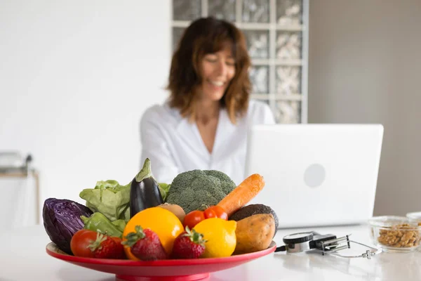 Female nutritionist working with laptop in a doctors office with big fresh vegetable and fruit plate. Concept of a healthy lifestyle, body care and weight loss. Selective focus. Blurred background.