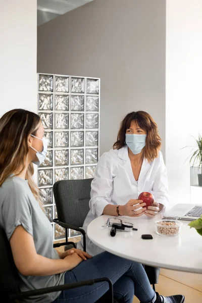 Dietitian with red apple speaking with woman, wearing protective face mask, while looking at each other at table with healthy ingredients, in a fashionable office. Vertical, copy space