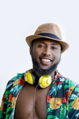 Black smiling man in cool flower shirt and had, with headphones on neck looking at camera while standing against white background. Vertical portrait