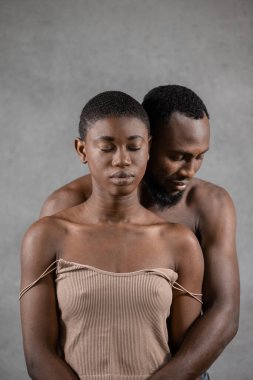 Shirtless Black man embracing from the back his girlfriend with closed eyes against gray background. Vertical copy space