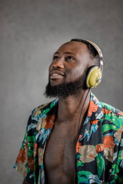 Smiling Black man wearing flowery unbuttoned shirt and headphones standing sideways looking up against gray background. Vertical portrait