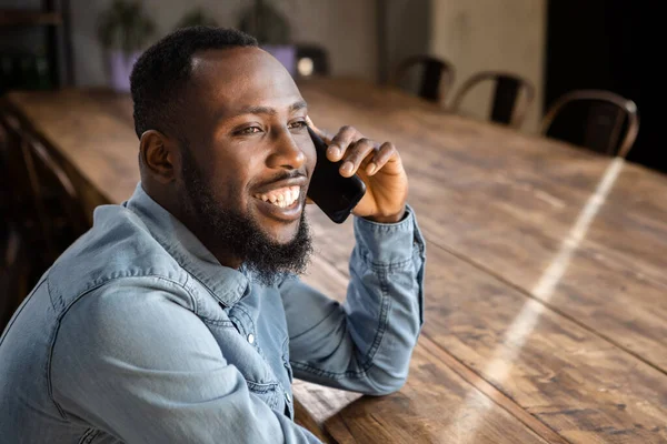 Smiling Young Black man in denim shirt sitting at big office table having phone call looking away. Horizontal copy space