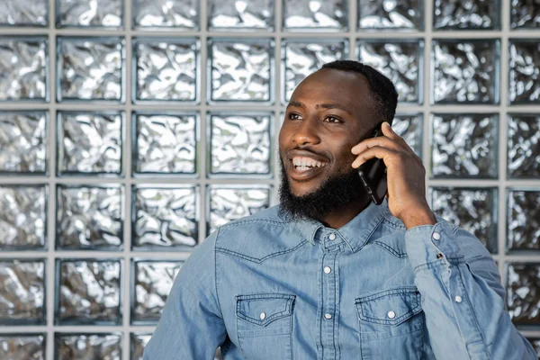 Smiling Young Black Afro man wearing jeans shirt having phone call looking away against glass block wall in a fashionable office on daytime. Copy space, horizontal