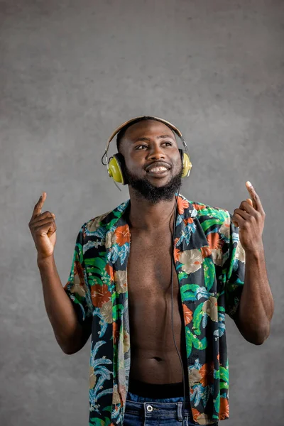 Young smiling Black man dancing while listening to music in headphones against gray background. Vertical portrait, copy space.