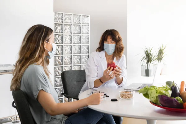 Dietitian with red apple speaking with woman in sterile mask while looking at each other at table with healthy ingredients. Wide angle, horizontal