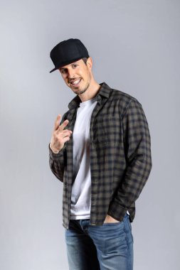 Gri arka planda izole edilmiş, beyaz tişörtlü, kareli tişörtlü ve şapkalı parmaklarını zafer işareti yaparken gösteren kameraya bakan mutlu genç bir adam. Dikey
