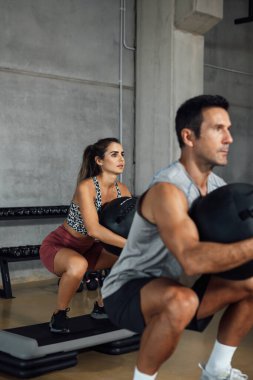 Sporty man and woman doing squats holding fitness ball in the gym. Training legs and core ab workout. Sport, training and lifestyle concept. Vertical, copy-space.