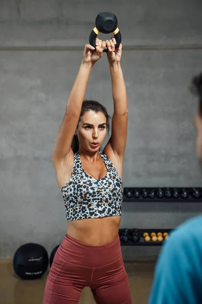 Fit caucasian Athletic woman lifting weight above his head with two hands, doing kettle bell or dumbbell workout in dark gym, looking at instructor. Vertical