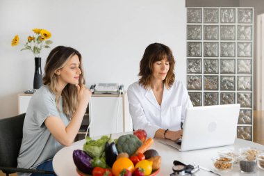 Dietitian typing on netbook and talking to content woman while looking at screen at desk with healthy food indoors. Copy space