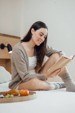 Serene brunette young woman reading book on soft bed with breakfast on tray, in the morning, daylight. Vertical.