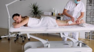 Woman on table against unrecognizable beautician in uniform and sterile mask with waxing strip during hair removal process on table. Video horizontal hd 4k, real time.