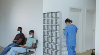 Siyah kadın ve erkek aşıyı almadan önce bekleme odasında cep telefonu kullanıyor. Koruyucu üniformalı kadın doktor bir sonraki hastayı aşı için arıyor, gerçek zamanlı, 4K video..