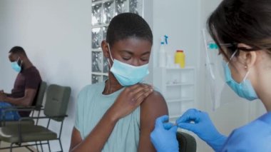 Unrecognizable female nurse in protective uniform, latex gloves vaccinates and puts a band-aid on African American female patients arm in clinic office during coronavirus outbreak. 4k video