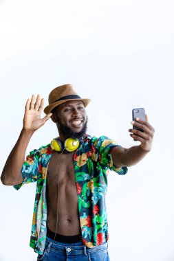 Young Smiling Black man with flowery unbuttoned shirt and headphones on neck standing waving hand while taking selfie against white background. Vertical Copy space