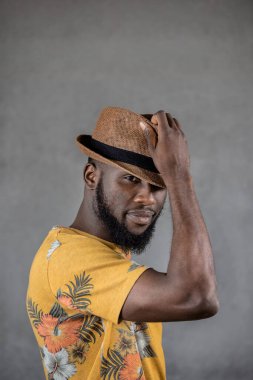Bearded young black man in flowery yellow t-shirt touching hat as he stands sideways against gray background. Vertical copy space.