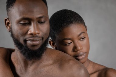 Serious Black woman embracing Black shirtless man looking down against gray background. Horizontal