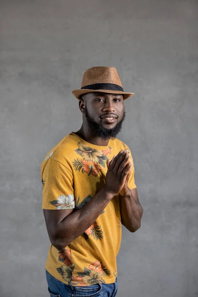 Smiling black man in hat and yellow flowery t-shirt standing with hands together, prayer gesture, looking at camera against gray background. Vertical