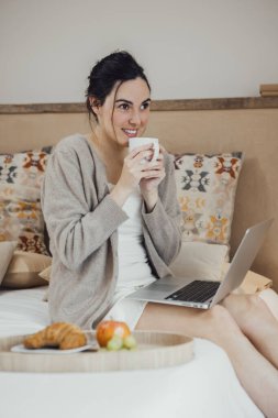 Smiling Young brunette woman sits on bed with a mug on hand, using laptop and looking aside, morning daylight. Vertical