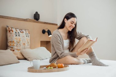 Serene brunette young woman reading book on soft bed with breakfast on tray, in the morning, daylight. Horizontal copy space.