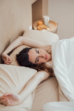 Portrait of young Serene woman waking up, lying on bed stretching arms after good sleep on bed, looking at camera with sleepy look. Horizontal