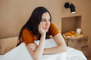 Sleepy dreamy woman sitting on a comfortable bed in the morning, looking away thoughtfully, resting her head with one hand in a cozy trendy bedroom. Earthy colors, Horizontal