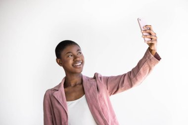 Smiling Black Young woman with buzz cut hairstyle in pink blazer taking selfie using smartphone while standing against white wall. Horizontal