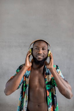 Young smiling Black man with hands on headphones looking at camera while listening to music in headphones against gray background. Vertical portrait, copy space.