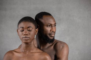 Shirtless Black man looking away embracing from the back his girlfriend with closed eyes against gray background. Horizontal copy space