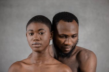 Shirtless Black man looking down embracing from the back his girlfriend who is looking at camera against gray background. Horizontal copy space