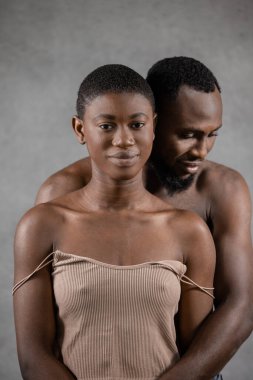 Shirtless Black man embracing from the back his smiling girlfriend with closed eyes against gray background. Vertical copy space