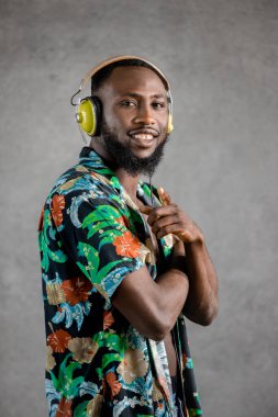Smiling Black man wearing flowery unbuttoned shirt and headphones standing sideways pointing and looking at camera against gray background. Vertical portrait