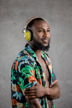 Smiling Black man wearing flowery unbuttoned shirt and headphones standing sideways looking away against gray background. Vertical portrait