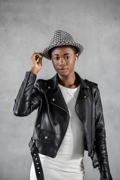 Portrait gorgeous Young Black woman wearing black leather jacket and white dress and short hair touching hat looking at camera against gray background. Vertical
