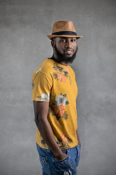 Bearded young black man looking at camera standing sideways with hands in pockets in flowery yellow t-shirt against gray background. Vertical