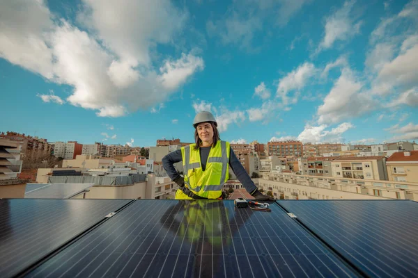Confident Female electrician engineer in safety helmet and yellow vest, looking at camera, by urban Solar Panels on a city rooftop, with blue sky background. Gender equality concept. Horizontal