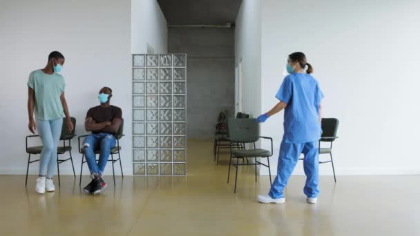 ワクチンを服用する前に携帯電話を使用して病院の待合室で黒人女性と男性 予防接種のために次の患者を呼び出す保護制服の女性医師 リアルタイム 4Kビデオ — ストック動画