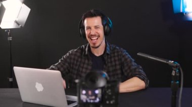 Genç beyaz adam dizüstü bilgisayarı ve kulaklığı ile oturuyor, kamerayla bir video blogu kaydediyor sosyal medya için canlı yayın yapıyor. Yatay, 4k video.