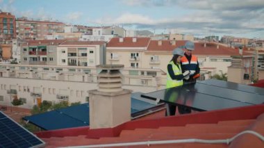 Güneş panelleri bakımı ya da fotovoltaik çatı teftişinde çalışan teknik mühendisler. Kadın, erkek işçiye yelek ve şapka kullanma talimatı veriyor. Yan görüş, uzak çekim, 4k yatay görüntü