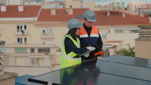 在光伏屋顶上从事太阳能电池板维修或检查工作的技术工程师 妇女向穿着背心和硬礼帽的男工发出指示 侧视图 4K视频水平 — 图库视频影像