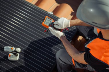 Güvenlik kaskı ve turuncu yelek giyen erkek mühendis şehrin çatısında dijital multimetre ile güneş panelleri voltajını test ediyor. Yenilenebilir enerji kavramı. Yatay, kapat, üst görünüm, boşluğu kopyala