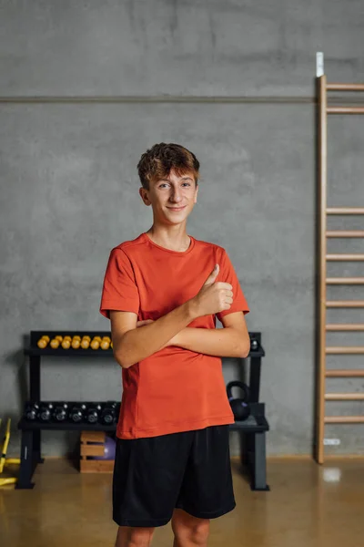 stock image Smiling and positive teenager shows thumbs up and looks at camera at gym, with concrete gray wall background and wooden wall bars. Sport, fitness. Portrait, copy-space, vertical