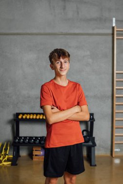 Gülümseyen ve kırmızı tişörtlü pozitif bir gencin portresi jimnastik salonunda kameraya bakarak, beton duvar arkası ve ahşap parmaklıklar. Spor ve spor. kopyalama alanı, dikey