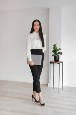 Kendine güvenen, beyaz, uzun saçlı, boş bir ofiste dizüstü bilgisayarla kameraya bakan zarif bir kadın. Yönetici, iş kadını. Tam uzunluk, Dikey.