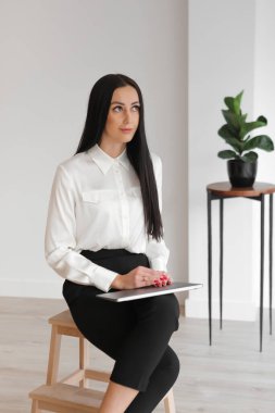 Uzun saçlı, düşünceli, zarif, yönetici bir kadının portresi. Taburede oturuyor, dizüstü bilgisayarını tutuyor, işyerine bakıyor. Dikey.