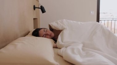Genç esmer kadın rahat bir yatakta huzur içinde uyuyor, uyanıyor ve sabah bir yatak odasında toprak renklerle esniyor. Yatay, hd video 4k çözünürlüğü.